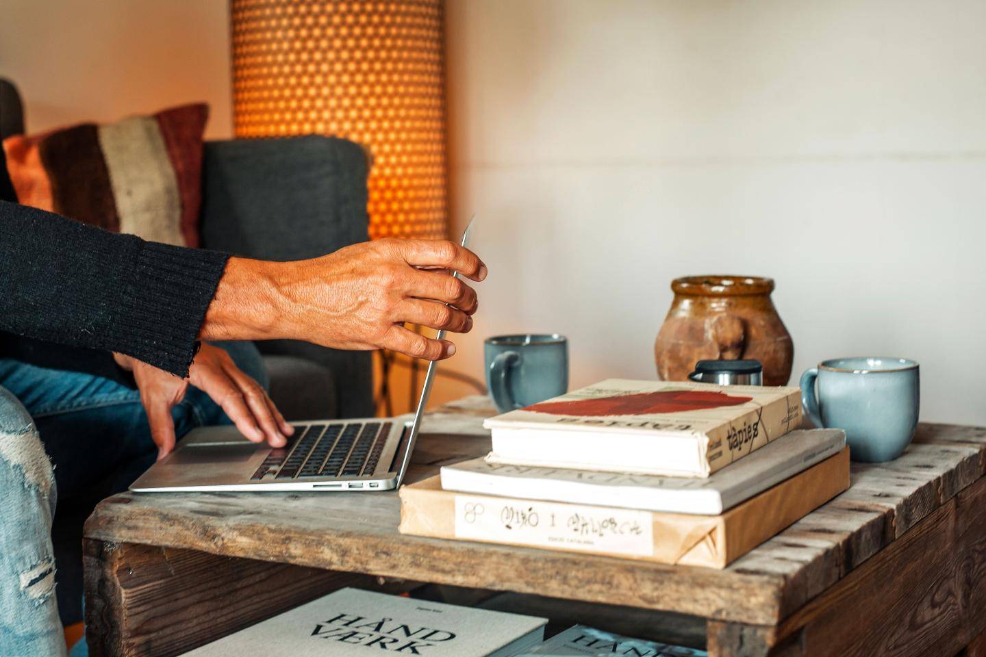 En person arbejder på en bærbar computer ved et lavt træbord med bøger, kopper og en krukke.