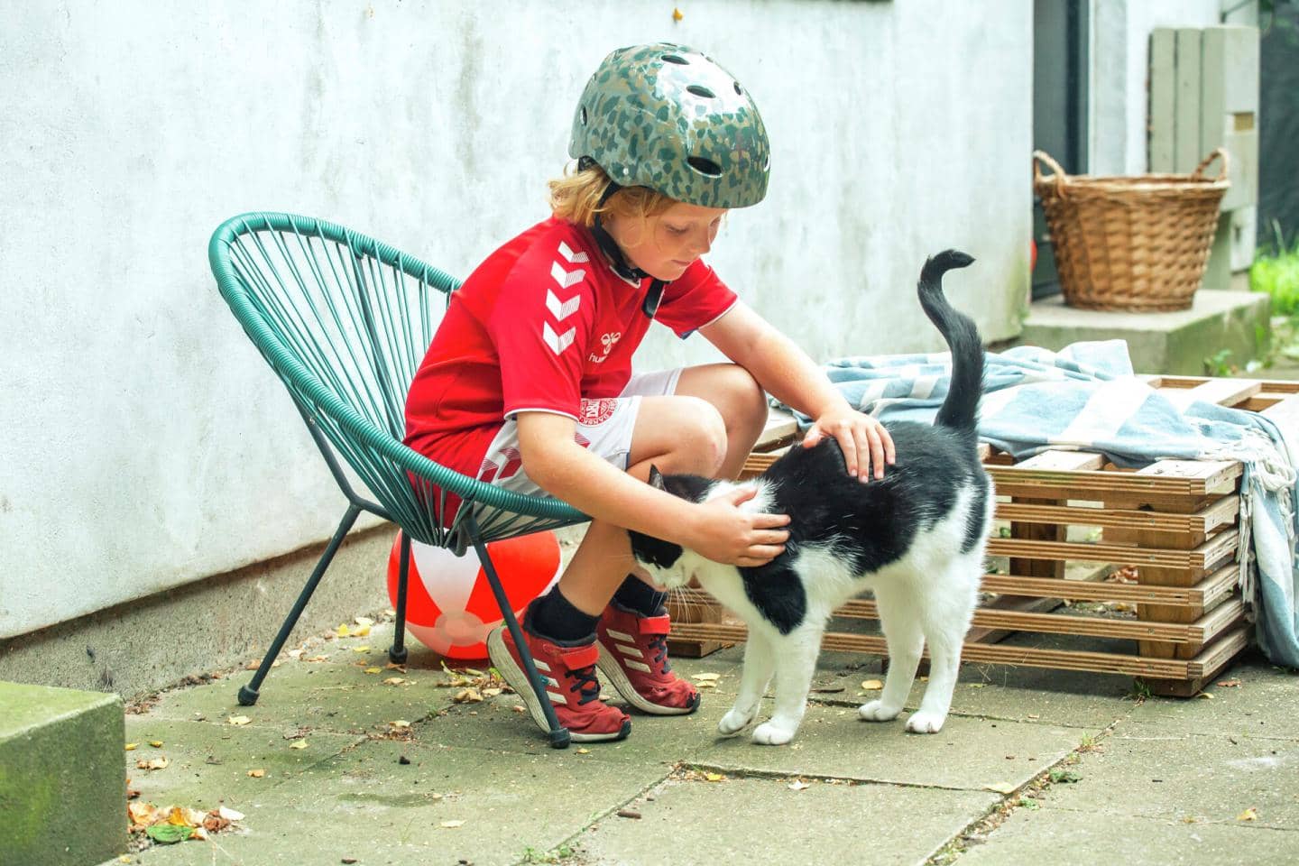 Dreng med cykelhjelm sidder i havestol og aer kat