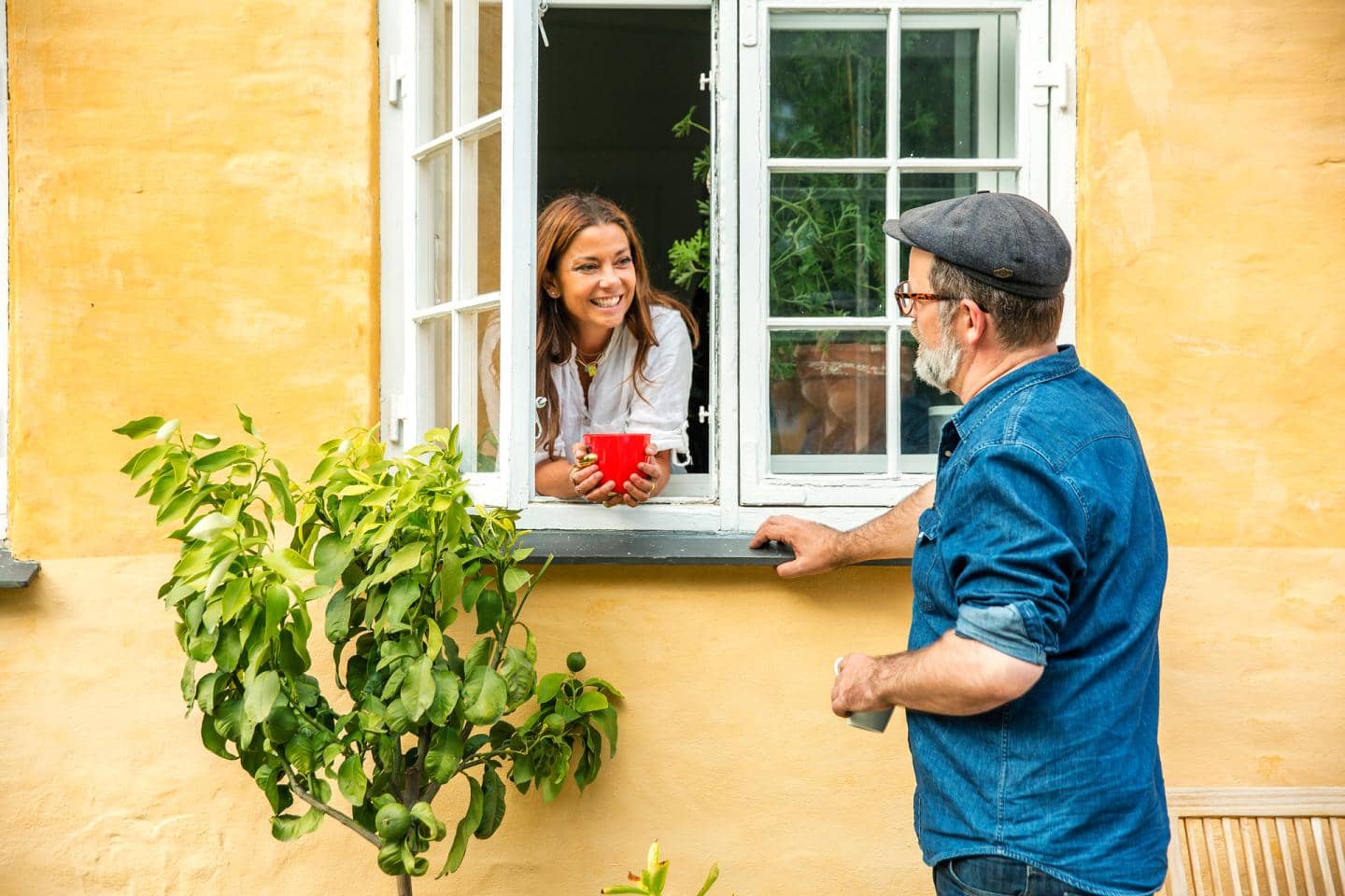 Kvinde læner sig ud af vindue på gult hus og snakker med mand, der står ved siden af vinduet. De smiler.