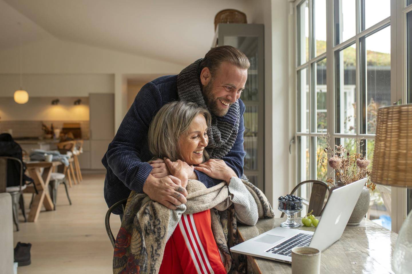 Kvinde sidder ved bord, mand krammer hende og kigger på computer på bord, fotografi