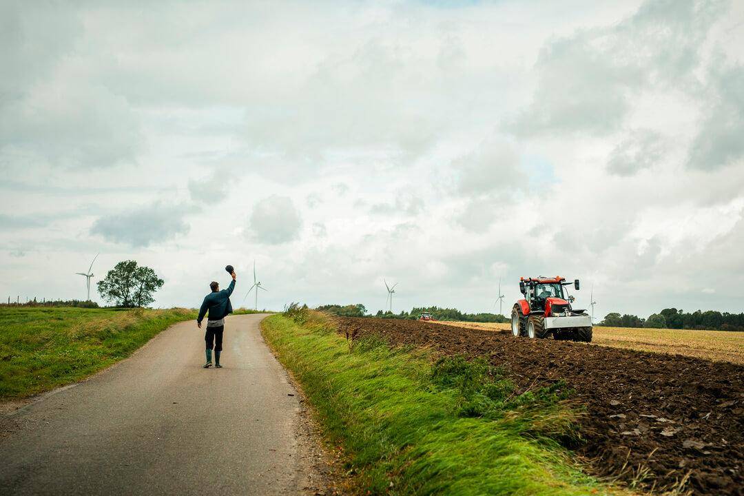 En mand set bagfra går langs en landevej og løfter sin kasket mod en rød traktor, der arbejder på en pløjet mark under en stor, skyet himmel med vindmøller i baggrunden.