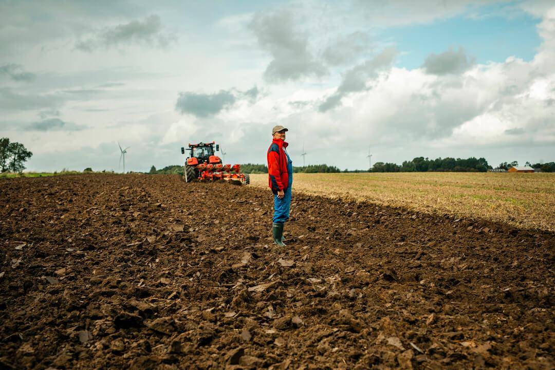 En landmand i blå overalls og rød jakke står på en pløjet mark, mens en rød traktor arbejder i baggrunden under en skyet himmel.